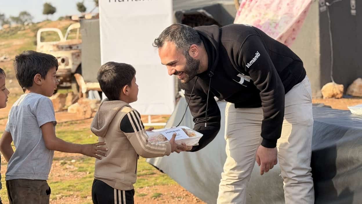 Aid worker handing food to displaced children near tent camp.